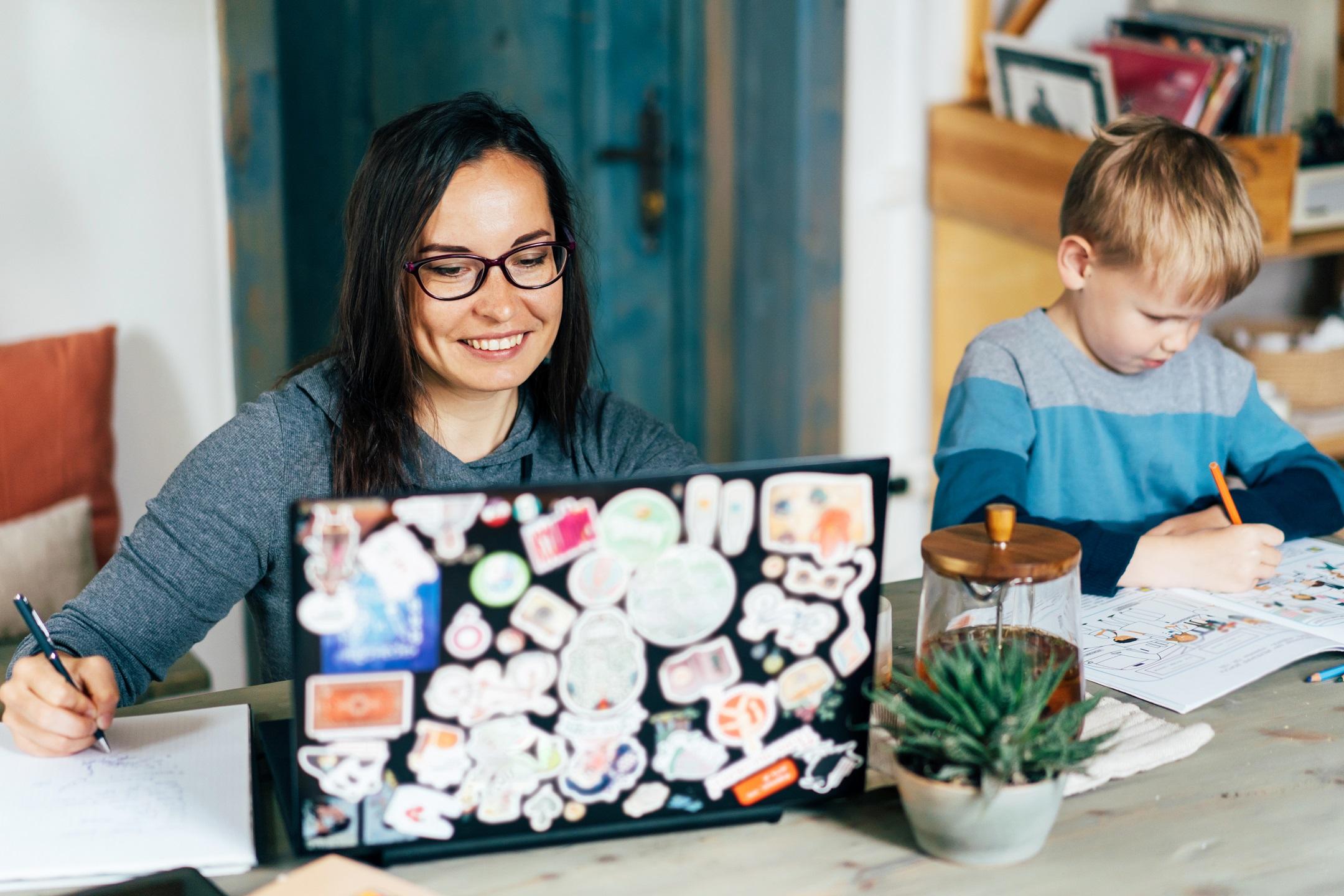 Eine Frau und ein junges Kind sitzen an einem Tisch, die Frau arbeitet an einem Laptop, während das Kind daneben in einem Malbuch malt.