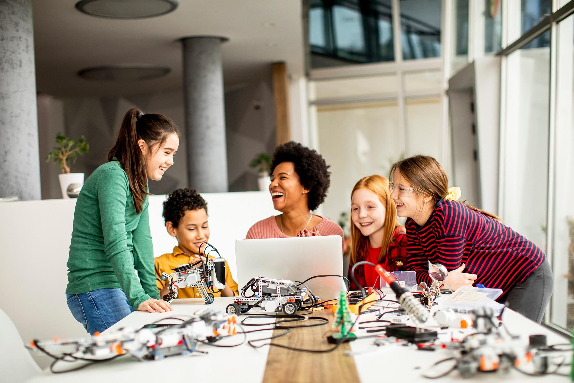 Glückliche Kinder mit ihrer afroamerikanischen Lehrerin für Naturwissenschaften mit Laptop, die elektrisches Spielzeug und Roboter im Robotik-Klassenzimmer programmiert.