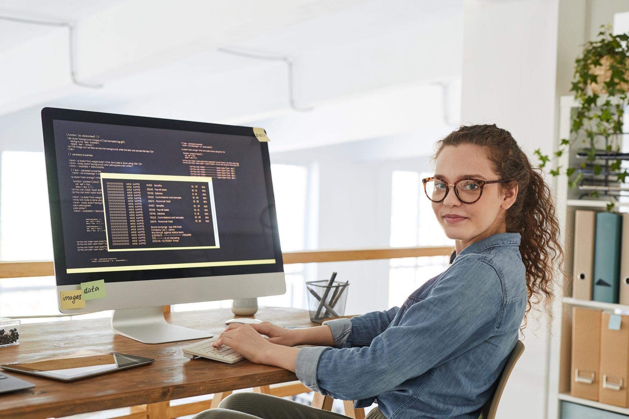 Eine junge Frau sitzt am Schreibtisch und dreht sich mit dem Kopf zur Kamera. Vor ihr auf dem Computerbildschirm ist Computercode auf schwarzem Grund zu sehen.
