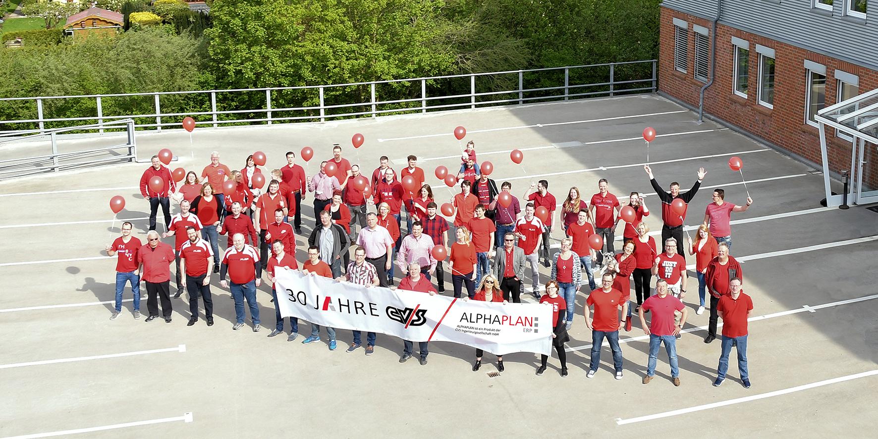 Eine große Gruppe Menschen steht draußen auf einem Platz und hält ein Banner. Das Foto ist aus der Luft aufgenommen.