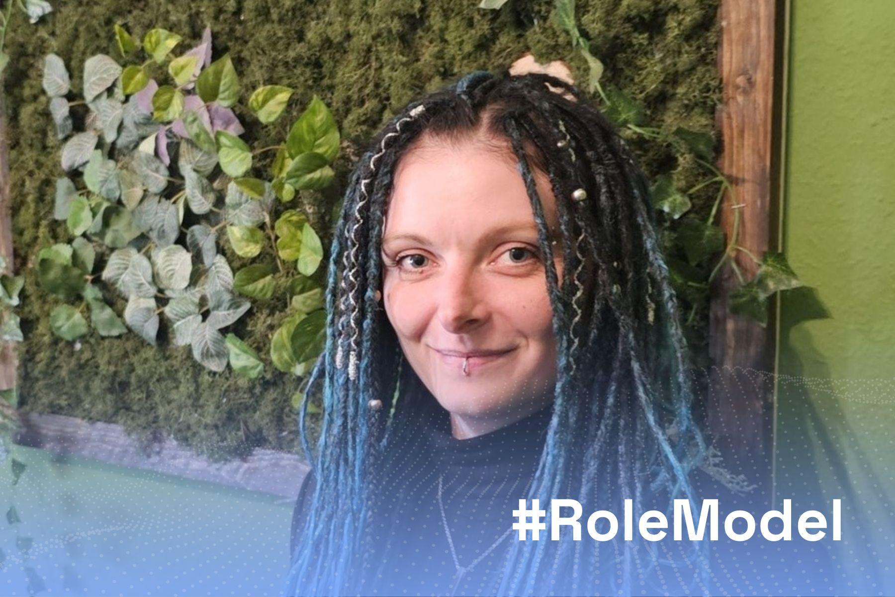Eine Frau steht vor einer grünen Wand und lächelt in die Kamera, darüber steht in weißer Schrift #RoleModel